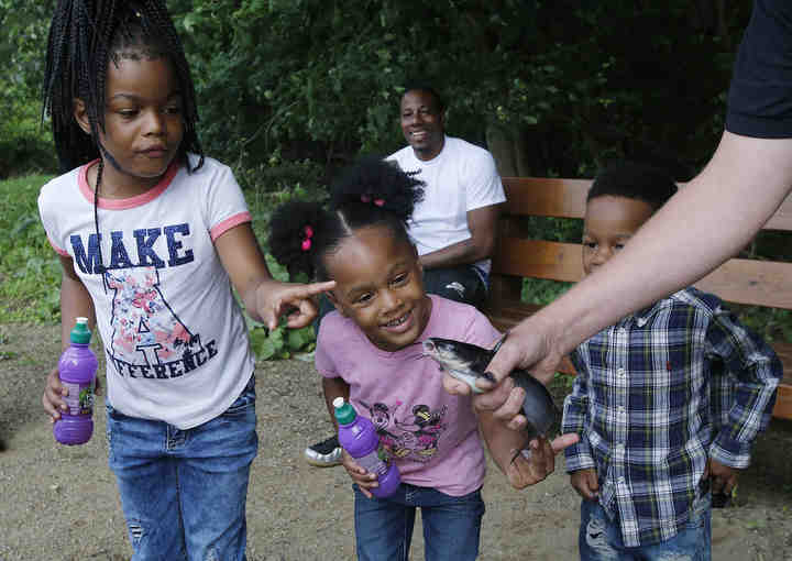 Darius Mitchell (seated background) of Akron smiles as he watches his children Tashayla Mitchell, 7, Darshay, 4, and Darius, 3, check out a catfish held by Jeromy Applegate, a biologist with the U.S. Fish and Wildlife Service as Applegate helps stock Alder Pond with 149 catfish in Akron.  (Karen Schiely / Akron Beacon Journal)